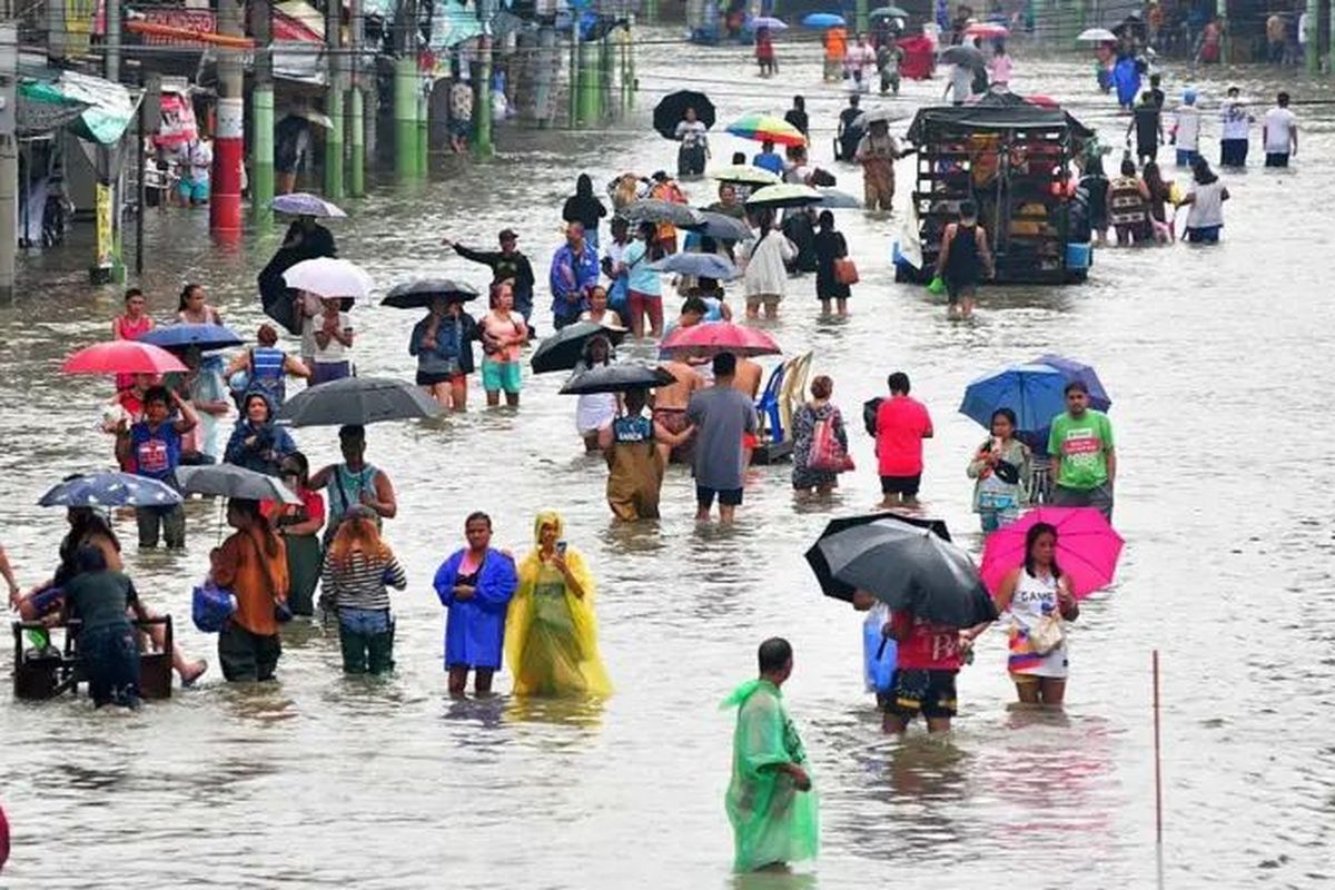 Puluhan orang memegang payung dan mengenakan jas hujan saat menerobos banjir di Filipina.
